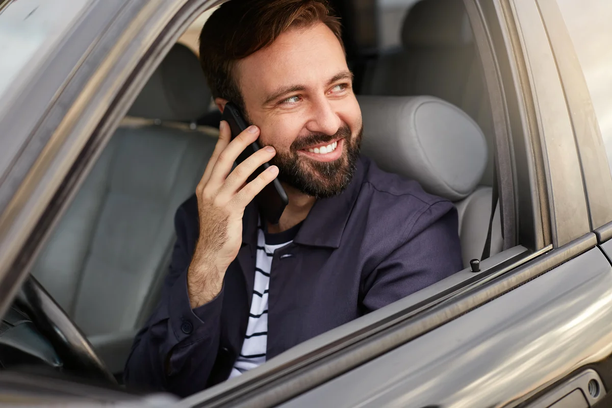 young happy attractive successful bearded man sits car talking phone with his friend smiling broadly looking away min